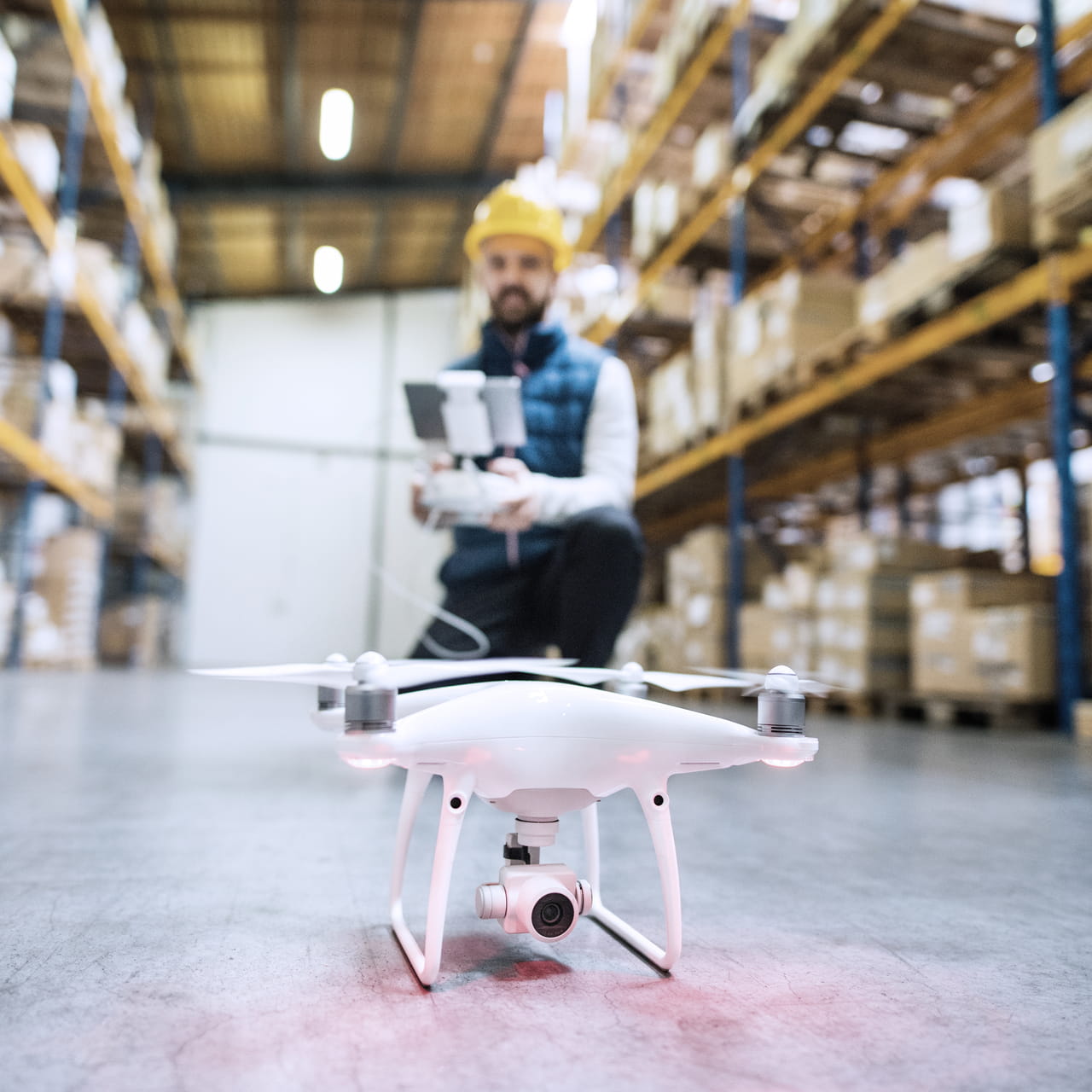 Drone on the floor of a warehouse. In the background is a man wearing a hard hat and a vest, operating the drone with a remote control